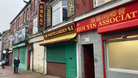 BBC /Claire Hamilton A row of terraced buildings with shops at the bottom, with shutters up on the front of the shops