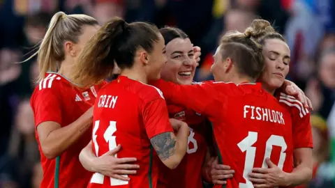 Wales' Angharad James celebrates scoring a second goal with teammates. She is smiling and being hugged by four other players in their red Wales shirts, including Fishlock and Cain. The background is blurred. 
