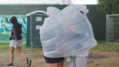 PA/ Yui Mok Two people standing near a green portaloo at the Glastonbury Festival, with a white plastic poncho stretched over both of them.