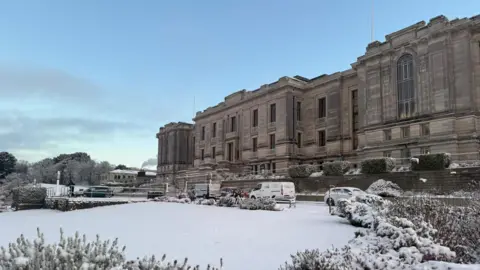 National Library of Wales Outside the National Library of Wales covered in snow. It is a grand stone building with large stone columns on the façade. Snow covers the garden in front of the building with some snow-coloured bushes in the foreground. 