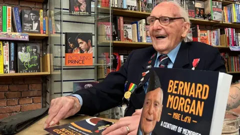 An older man, with numerous badges and medals on his blazer, is sat at a desk, smiling. In front and to the side of him are copies of his book, called Bernard Morgan,  A Century of Memories