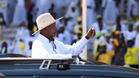 Uganda's President Yoweri Museveni, in a white shirt and a white hat, salutes his supporters from an open-top black vehicle after his swearing-in ceremony - 12 May 2026