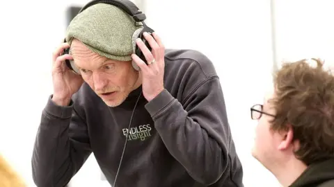 Mark Douet Paul Jenkins stands in a crouched position with hands over a pair of large earphones, with a look of concentration on his face. A second unnamed person's face is in the bottom right corner looking away from the camera towards Paul.