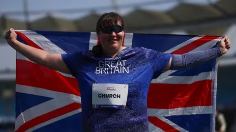 Getty Images/Dean Mouhtaropoulos Lydia Church sorri para a câmera e segura uma bandeira da Union Jack atrás dela. Ele usa óculos escuros e blusa azul com palavras 