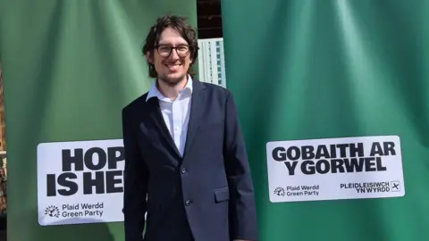 Angela Karadog A man in a navy suit and a white shirt, with dark hair and glasses stands smiling to camera. Behind him are two green banners with the Green party logo on. 