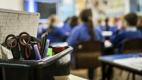 A box of stationary - including a whiteboard, pens and scissors - with a class of primary school students in the background.