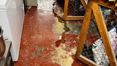 Phil Hall A trickle of water is running across a floor. The floor looks like rough concrete with orange paint. 