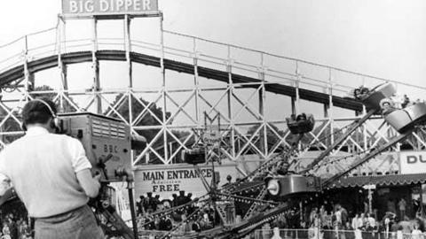 Black and white image of big dipper fair ride in the distance. Cameraman is visible in the foreground. Another ride swirls in front of the big dipper.  A sign is visible for the 'Main Entrance' and a sign for 'BIG DIPPER' is seen above the ride itself.