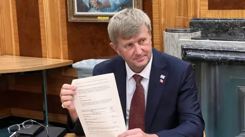 A man in a blue suit and red tie with an Essex lapel badge and union jack badge. He's holding up a piece of A4 paper with his signature in the middle.