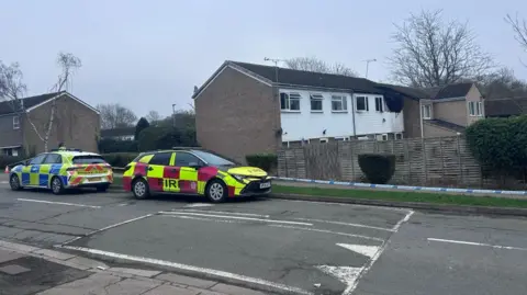 Kate Bradbrook/BBC A fire car and police car on a residential street. Behind them is police tape. Behind that are houses, one of which is fire damaged.
