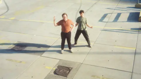 Joshua Jarman Josh and Ben standing on concrete both in dark trousers. Ben is in an orange top and waving one arm and Josh is in a green t shirt and holding his arms apart. 