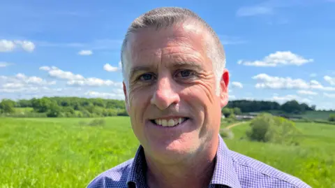 Paul is wearing a purple and white chequered shirt. He has short cropped grey hair. In the background are green fields and blue sky.