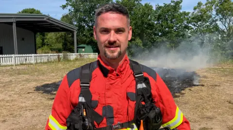 Bacup crew manager and burn team member Dean Barlow is in his uniform in front of a piece of scorched grassland.