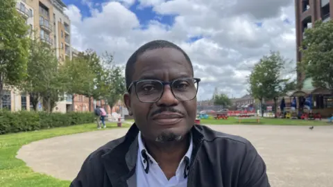 BBC Israel Eguaogie, wearing black glasses and a black jacket, sits in a public square in Belfast
