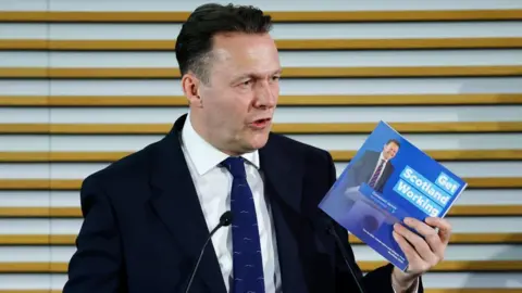 Russell Findlay, who has short black hair, speaks while holding a blue manifesto document. He is wearing a business suit and speaking into a microphone 