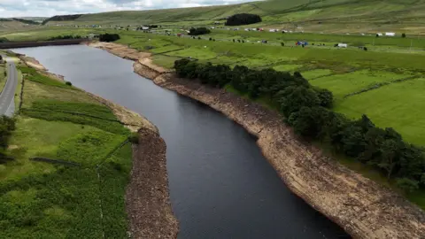Richard McCarthy/PA Wire Aerial view of a reservoir with visibly low water levels, exposing wide, dry, rocky banks. Surrounding the reservoir are green fields, scattered trees, and a road running parallel to the water.