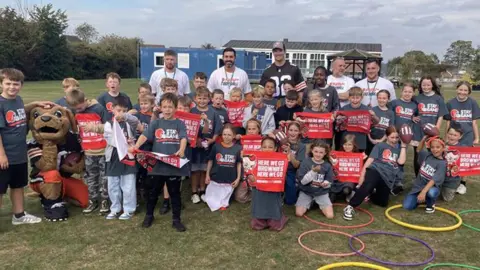 James Buriz/BBC é um enorme grupo de crianças do ensino fundamental na Thrapston Primary School. Eles estão usando camisetas cinzentas dizendo que ficam no jogo e seguram os banners vermelhos aqui vamos aos brownies aqui. Existem três ou cinco homens na camiseta atrás deles.