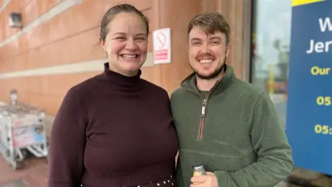 A couple smiling at the camera standing outside the arrivals area at Jersey Airport.