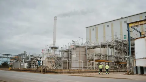 The titanium dioxide (TiO2) pigment production facility at Greatham, in Hartlepool. Two men in high viz jackets and hard hats walk towards industrial buildings. There is a large smoking chimney on the left, with tanks and several sets of external stairs visible on the right.