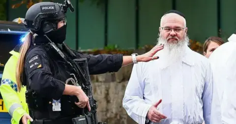 AFP via Getty Images Rabbi Daniel Walker, wearing a white kittel, is standing next to an armed police officer. 