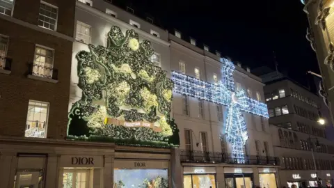 Dior window on New Bond Street at night with Christmas mural decorated with green and white lights and next to it a huge blue, purple and white bow lit up and wrapped around the building.