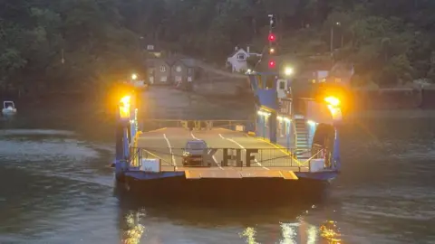Chain Ferry Challenge A generic view of a chain vehicle ferry in the dark. The large ferry is floating on top of the water. A car is parked on top of the vessel which is lit up. Houses are in the background as well as trees.
