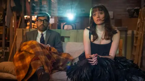 BBC/Gaumont/Ben Blackall Nabhaan Rizwan in a grey suit and Aimee Lou Wood in a black feathered dress, sitting on a bench watching a screen in a shed with a film projector emitting a beam of light behind them