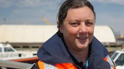 Woman in high-vis orange jacket smiling, looking at camera. Small white boat in background.