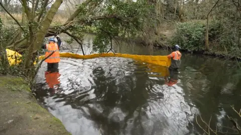 Two men in orange hi-viz and waders are in the river setting up a large yellow barrier to prevent the diesel spreading further downstream. 