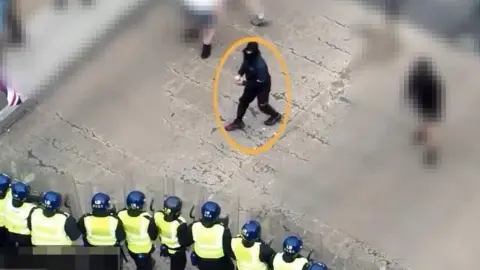 Northumbria Police Aerial view of a line of police officers confronting several men. Kennedy is dressed all in black with a black face covering and his holding a brick in preparation to throw it at the officers, who are pretty close to him.