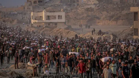 large crowds, some carrying sacks, walk past ruined buildings in Gaza