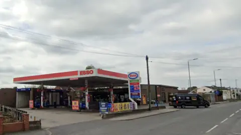 A picture of a petrol station forecourt with the word Esso above the station with a wall to the left of the forecourt and a brick building to the right of the petrol station