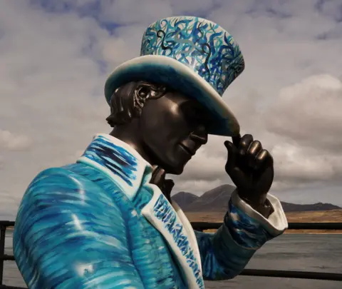 Sarah Sivers A view of a statue of a man, with a top hat and coloured blue and white. He has one hand touching the top of his hat, and through the gap in-between the crook of his arm and his head you can see three conical mountains.