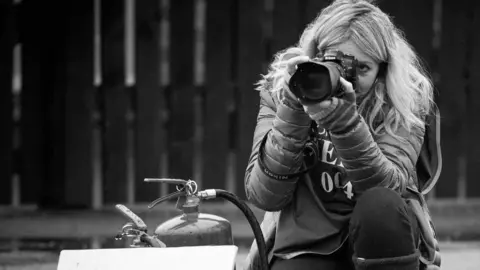 Jennifer Charlton Photographer Jennifer with long blonde hair and warm jacket and gloves peers through the lens in a black and white picture.