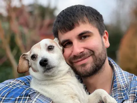 Man with brown hair and chequered shirt holding a small white dog with brown ears