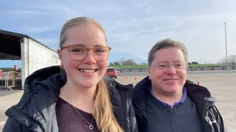 Woman in glasses smiling at the camera next to a man in glasses smiling at the camera in outdoor setting