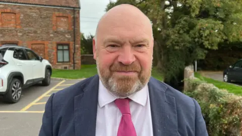 A man in a pink tie and blue jacket standing in a car park