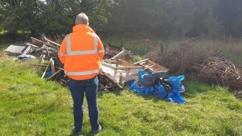 Wiltshire Council Image shows a man, with his back to the camera, in a florescent orange jacket looking at a pile of fly-tipped waste in a field. 
