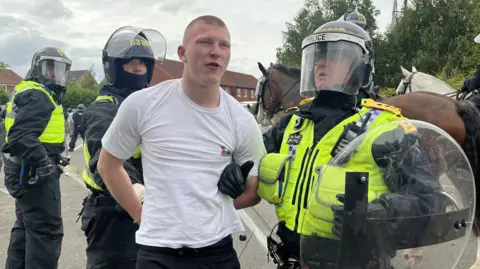 BBC News A man in a white t-shirt is led away by a police officer in riot gear