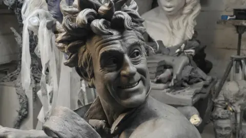 A close up picture of the clay model used for the sculpture showing Terry Jones with wild hair and smiling. He wears only a tie that looks like it is blowing in the wind