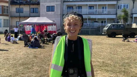 Kate Malyon wearing a beret and a high-vis jacket. An event is taking place behind her under a gazebo.