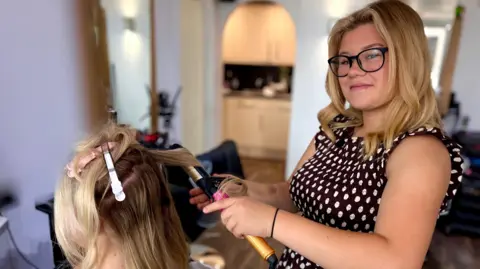 Martin Giles/BBC Jessica Flaxman stands in a hair salon, wearing a brown polka-dot dress and black-rimmed glasses, with her long blonde hair visible. She is using curling tongs on a client seated in front of her. The client, who also has blonde hair, has several styling clips sectioning her hair.