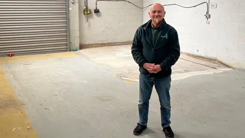 Worcester Foodbank A man wearing a dark-coloured jacket and jeans stands in an empty unit with a metal door and white brick walls.