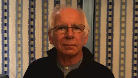 A balding man with grey hair and glasses looks at the camera. He is wearing a blue shirt and dark blue quarter zip, and stands against a pattered blue white wall.
