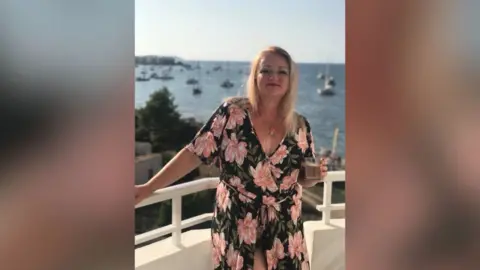 Gabby Rudge-Cox A woman in her forties is standing on a balcony in the sunshine. Behind her you can see the sea, which has yachts moored in a harbour. 