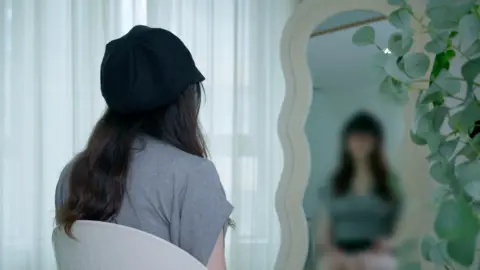 Back view of Baobao looking into a mirror, in which her image has been blurred to conceal her identity. She has long black hair and wears a grey T-shirt and black cap.