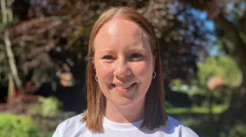 Sam Perkins's wife Emma Perkins, a white woman with short brown hair, wearing a white t-shirt and small hoop earrings.
