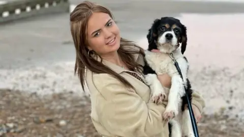 Hannah Byrne holds a small spaniel in her arms, both are looking at the camera. She is standing on a beach, with the waves seen behind her and the stones are visible on the beach.