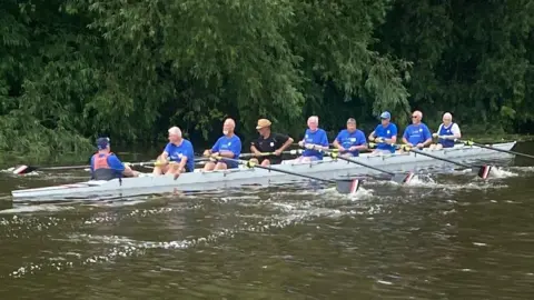 A group of eight men in blue t-shirts rowing. There is a cox at the back of the boat. 