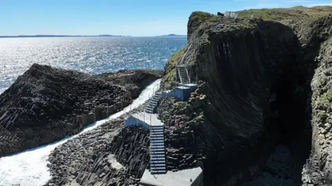 Staffa's new staircase in sunshine. The steps are made of concrete and go up the side of a steep sea cliff. Sunshine sparkles on the sea behind the island. 